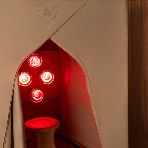 A FireLight Sauna against a bamboo wall, with the FireLight Bulbs emitting a warm red glow from within the organic cotton canvas inclosure.