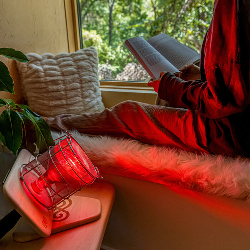 SaunaSpace Glow Infrared Therapy Light - A woman is lounging by the window reading her book while receiving warmth and red light therapy from the Glow.