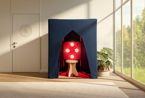 A compact booth with a dark blue cover, the SaunaSpace FireLight Infrared Sauna features EMF Shield, a wooden stool, and several red lights inside. It sits in a bright room with large windows and a nearby potted plant.