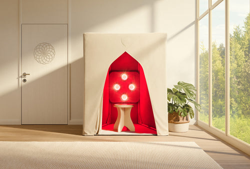 A compact beige EMF Shield tent by SaunaSpace, featuring a stool and four red-glowing FireLight Infrared Sauna lights inside, is set in a sunlit room with large windows and a green plant.
