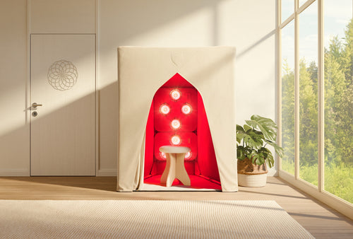 The SaunaSpace FireLight Infrared Sauna, beige and tent-like with an open front, displays a stool and red glowing lights inside, set in a bright room with large windows and a potted plant.