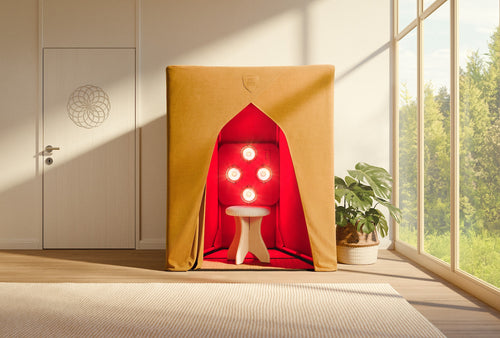 A compact, fabric-covered FireLight Infrared Sauna by SaunaSpace features red interior lighting, four circular lamps, and a white stool. The sauna is set in a bright room with large windows and a plant.