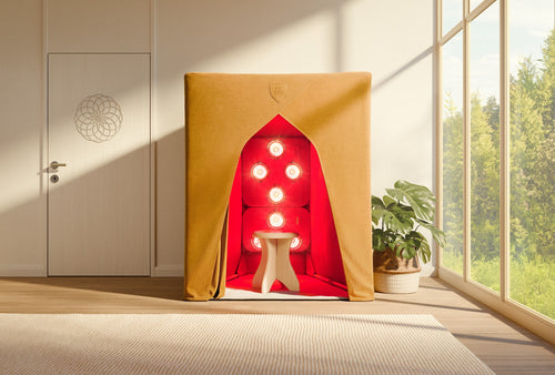 A compact SaunaSpace FireLight Infrared Sauna with glowing red lights and a white stool sits in a bright room with nearby greenery visible through large windows and a plant in the corner.