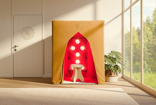 A tan fabric FireLight Infrared Sauna by SaunaSpace with red interior and six round lights on the back wall features a SilverLining™ EMF Shield and a white stool, placed beside a plant in a bright, modern room with large windows.