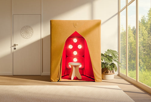 A tan fabric SaunaSpace FireLight Infrared Sauna booth with six glowing red lights and a white stool inside, set in a bright room with large windows, a plant, and a patterned door.