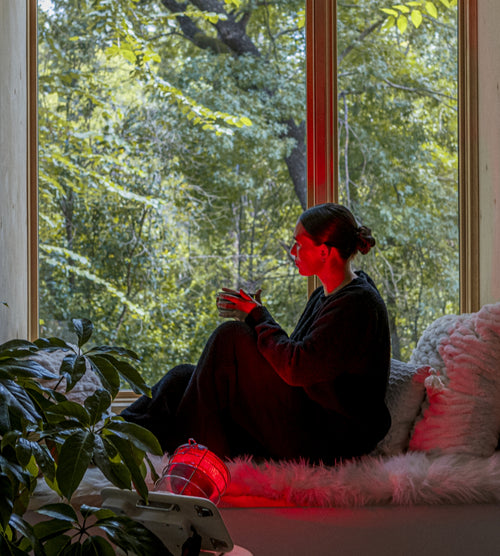 Woman seated against pillows on a sheepskin in sill of a large bay window drinking tea with Glow Infrared Therapy Light shining on her from side, next to a plant