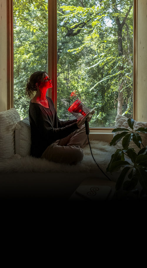 Woman shining Glow Infrared Therapy Light on face next to window with trees in background