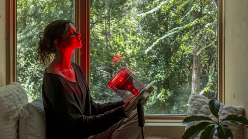 Woman shining Glow Infrared Therapy Light on face next to window with trees in background