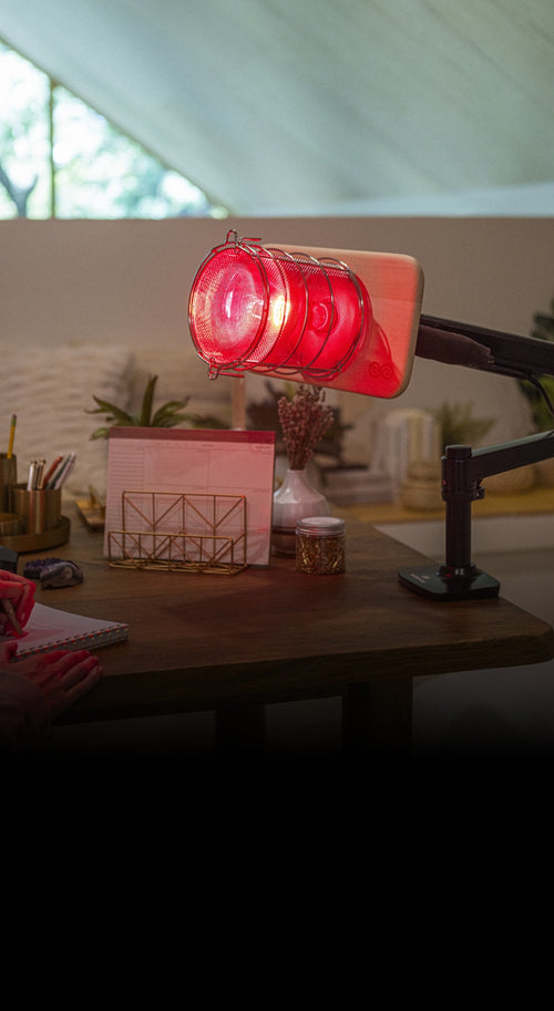 Glow Infrared Therapy Light mounted on Glow Desk Arm which is mounted on office desk with pens and papers and plants in background