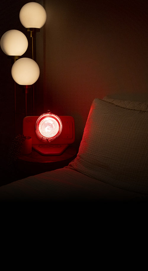 Glow Infrared Therapy Light turned on and rested on night stand next to bed with globe lights above in dimly lit bedroom