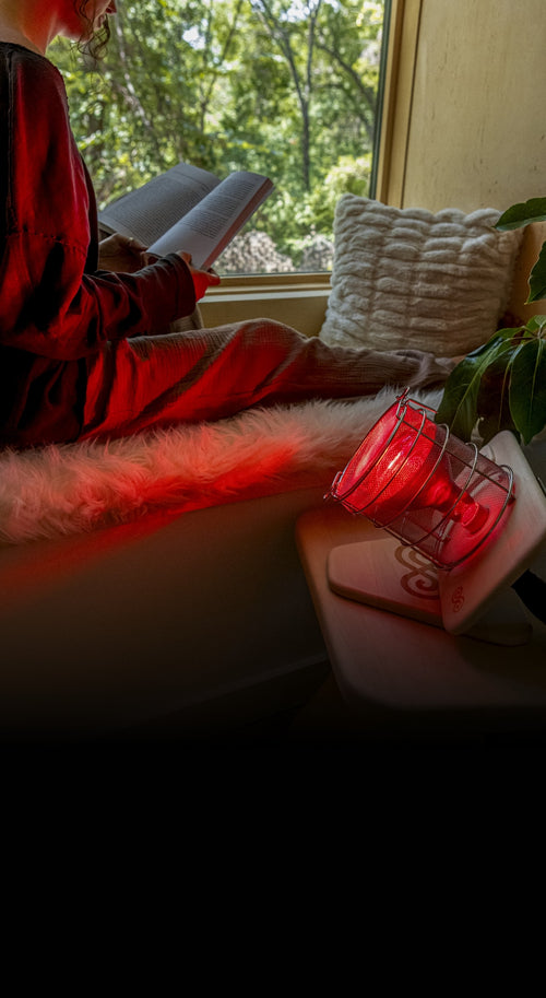 Woman reading book sitting in window sill with Glow Infrared Therapy Light on.