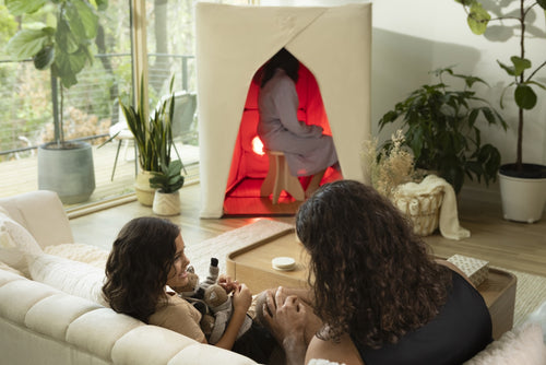 Family in living room with daughter and dad on couch and mother in FireLight Infrared Sauna in background, with plants and large windows.