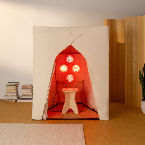 A FireLight Sauna set up in a bright living room with stacks of books on the left side. The FireLight Bulbs emit a warm, healing incandescent red light glow from within.
