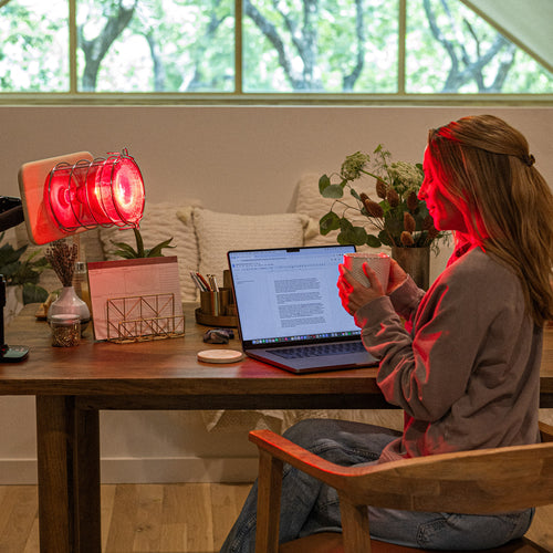 SaunaSpace Glow Infrared Therapy Light - Attached to a desk, shining on a woman while she works at her computer, counter-acting the harmful blue LED screen light.