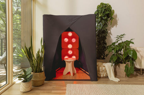 FireLight® Infrared SuperSauna in dark chocolate brown, illuminated with red light therapy bulbs, styled in a bright living room with greenery and woven accents.