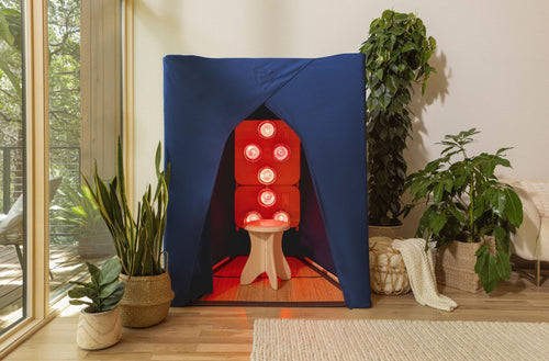 FireLight® Infrared SuperSauna in indigo blue, glowing with six red infrared bulbs inside a cozy modern living room with plants and natural light.