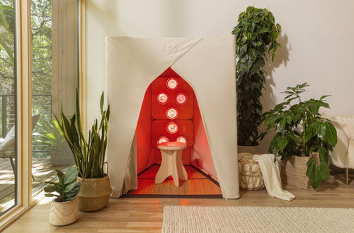 ireLight® Infrared SuperSauna in natural cream, lit with six red light therapy bulbs, featured in a modern living room with houseplants and woven textures.