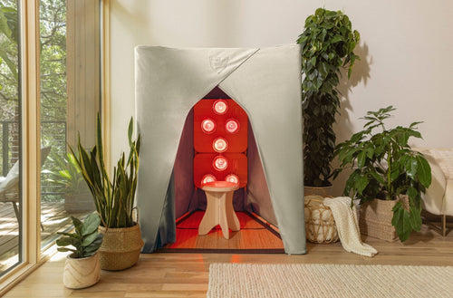 FireLight® Infrared SuperSauna in stone gray, glowing with infrared therapy bulbs inside, shown in a sunlit living room with plants and a cozy blanket basket.