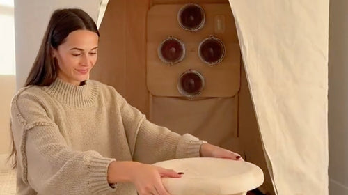 A woman in a beige knit sweater sits indoors holding a sauna stool next to a FireLight Infrared Sauna.