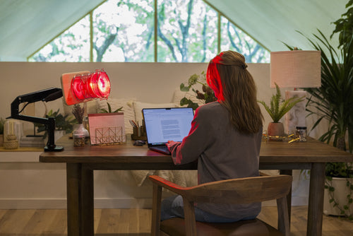 Woman sitting at desk with SaunaSpace Glow Desk Arm mounted on desk with Glow Infrared Therapy Light installed