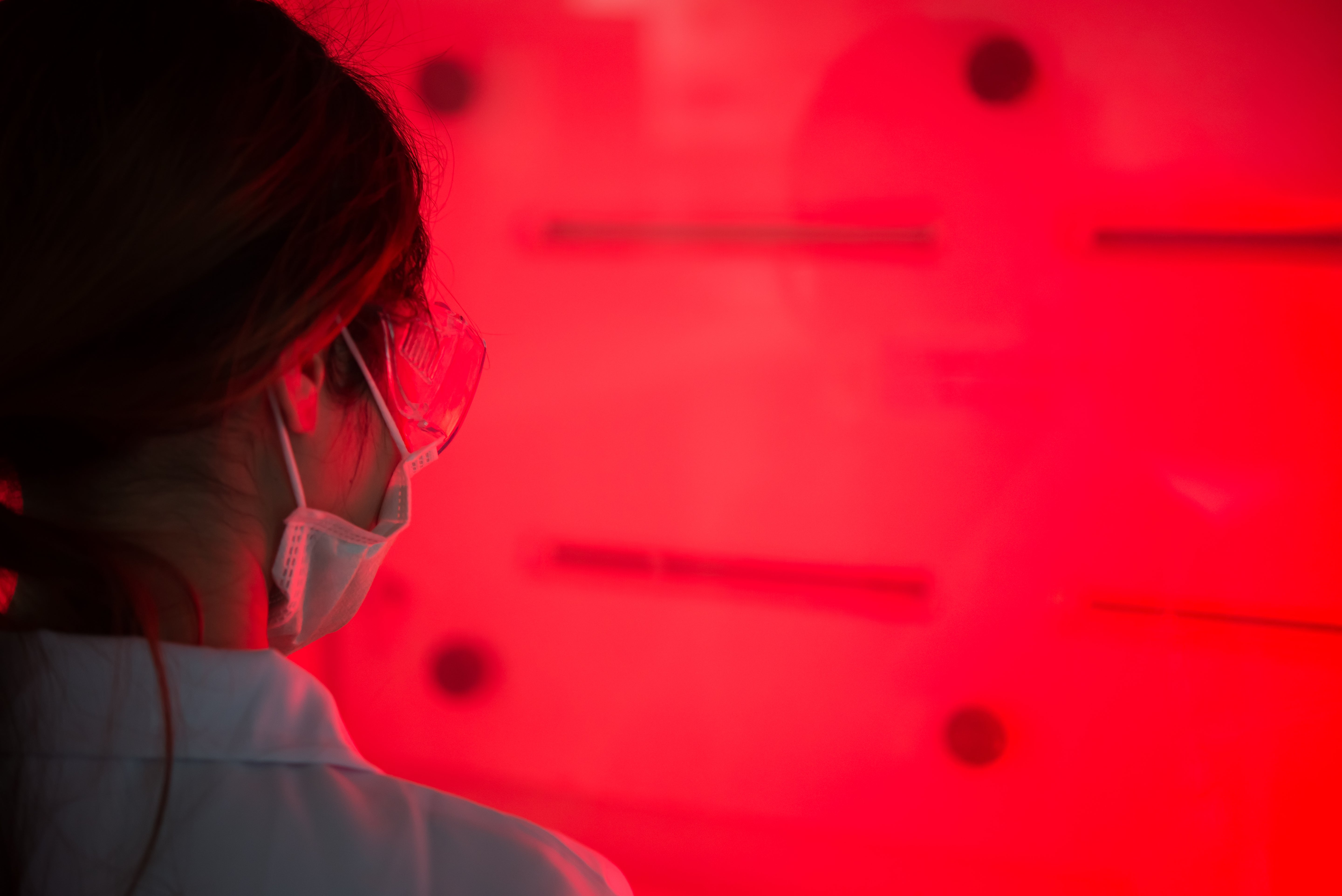 Woman in lab wearing safety glasses and mask doing research with red light in background