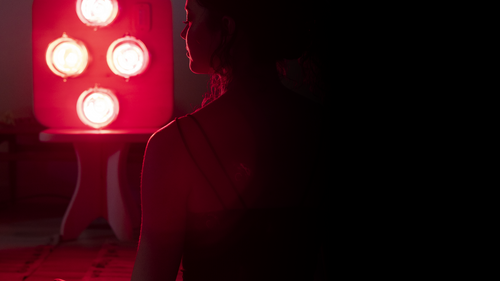 Silhouette of woman with eyes closed and seated in meditation in front of Hearth Quartet Infrared Therapy Panel with lights on that is resting on a Hearth Pedestal in dimly lit room