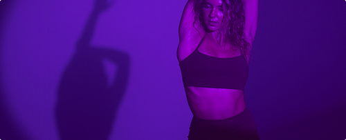 Woman dancing with arms raised with her shadow behind her under dark glow of violet light