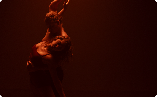 Man ballet dancing with and holding woman in dimly lit room with red light cast upon them