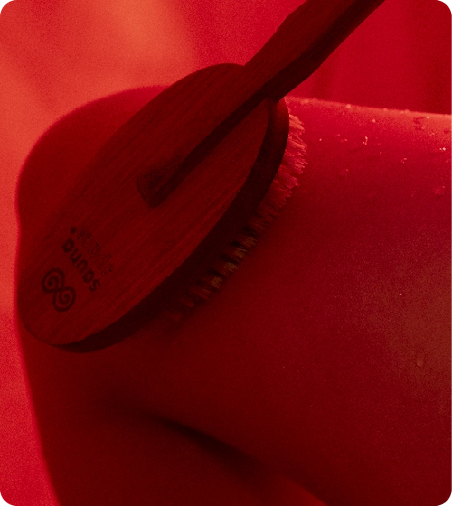 Woman using SaunaSpace Exfoliating Brush on her thigh under red light, inside FireLight Infrared Sauna