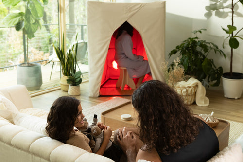 Family in living room with daughter and dad on couch and mother in FireLight Infrared Sauna in background, with plants and large windows.