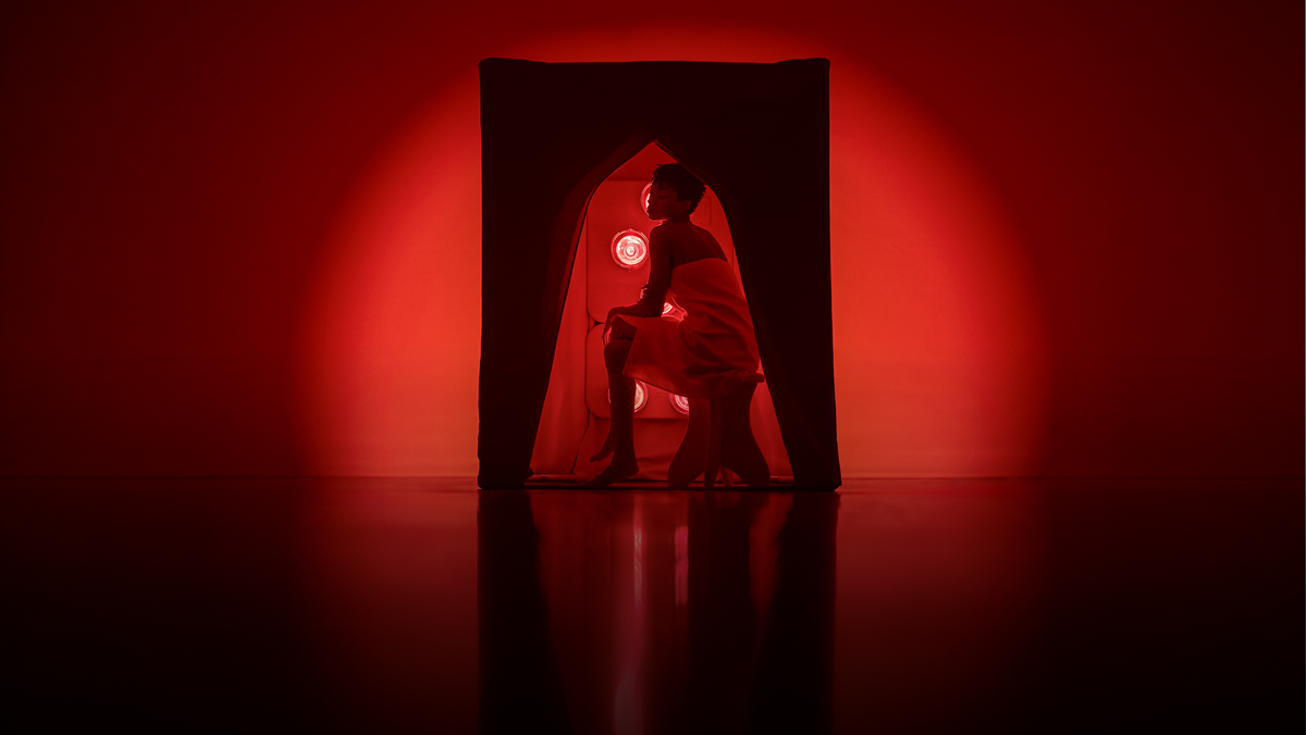A woman wrapped in a white towel sits inside a FireLight Infrared Sauna on a dark reflective floor, lit by dramatic red stage lighting and a red circular light in the background.