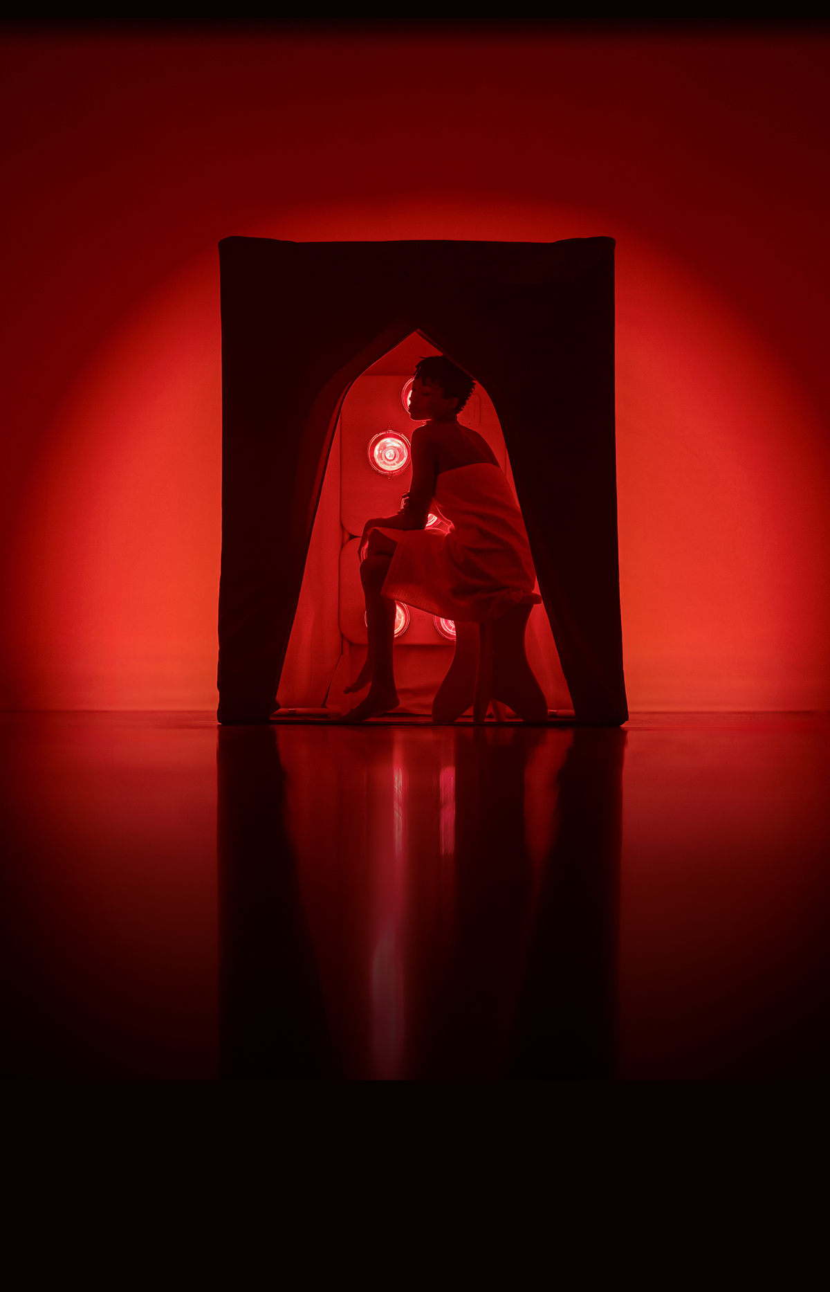 A woman wrapped in a white towel sits inside a FireLight Infrared Sauna on a dark reflective floor, lit by dramatic red stage lighting and a red circular light in the background.